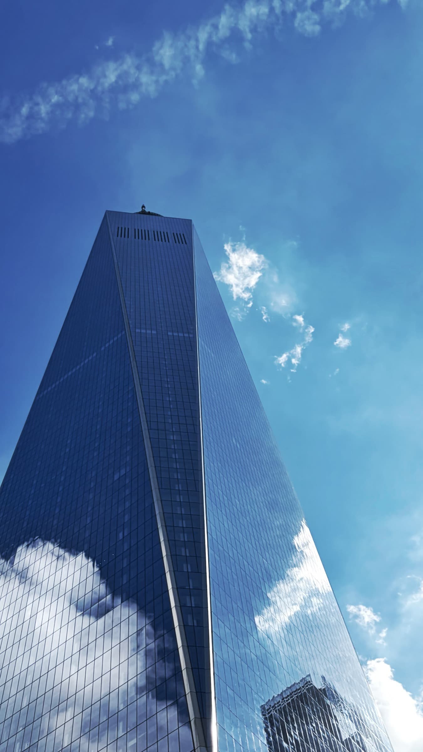 One World Trade Center photographed from below against a blue sky with cloud reflections